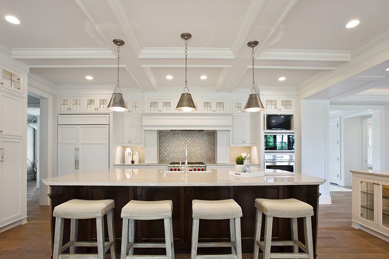 Well lit and all white aesthetic home kitchen with recessed ceiling lights, hanging lamps above the island, and lit backsplash